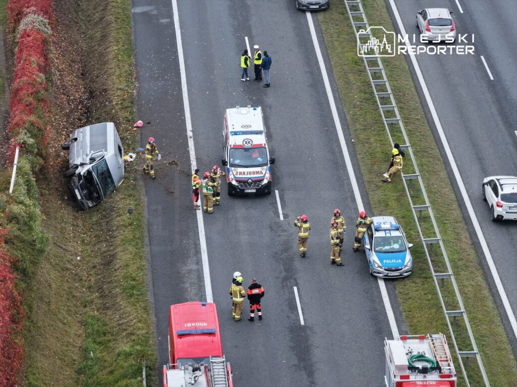 Karetka i radiowóz policji na poboczu drogi, obok przewrócony samochód oraz strażacy zabezpieczający miejsce wypadku.