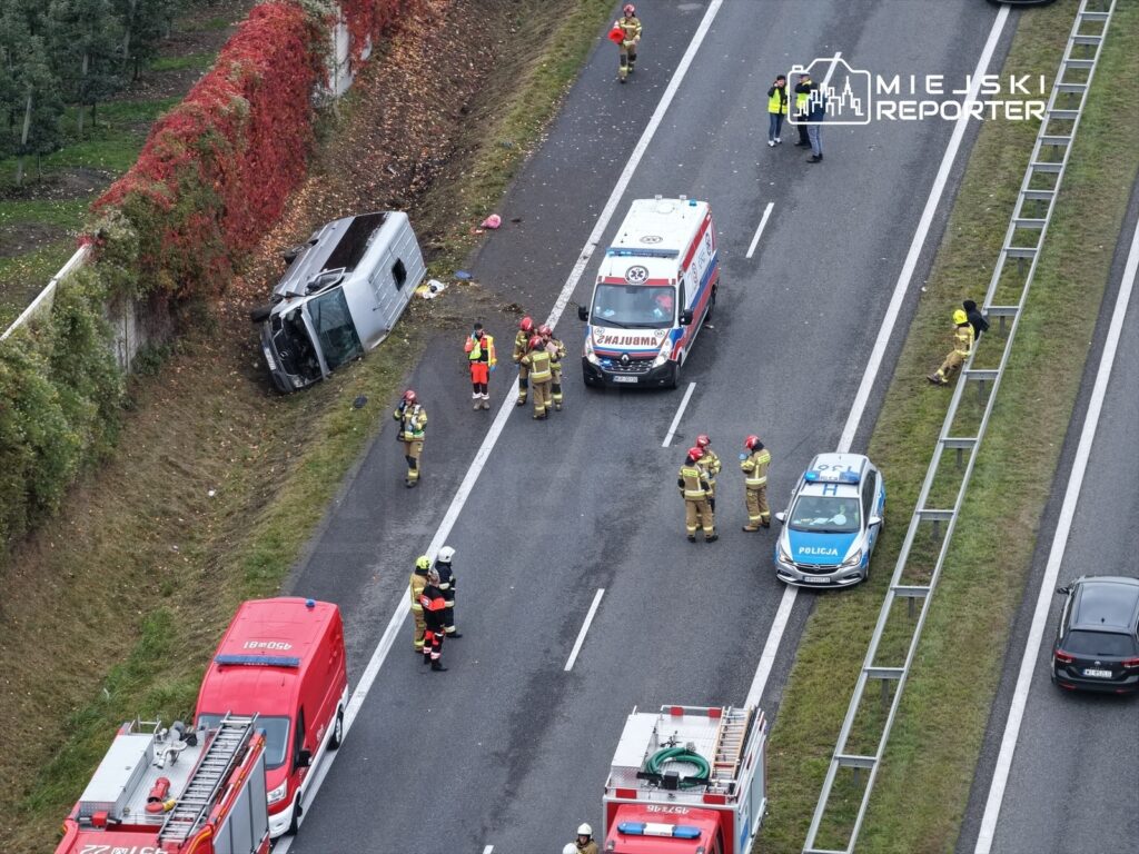Na drodze leży przewrócony pojazd, a obok grupka strażaków i ratowników medycznych udziela pomocy.
