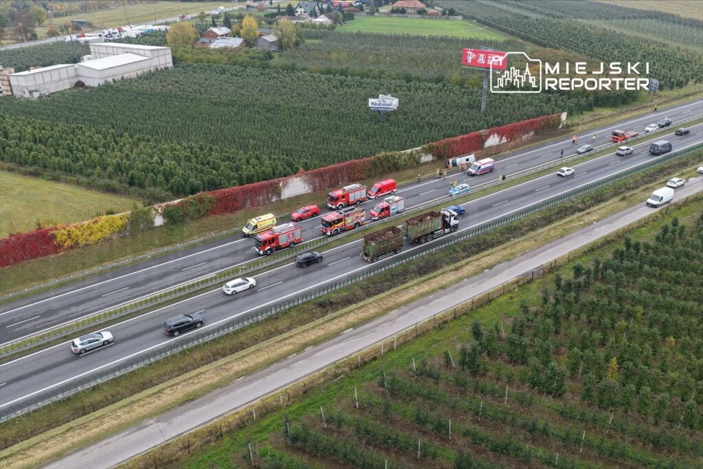 Na autostradzie zatrzymane pojazdy oraz pojazdy służb ratunkowych, w tle pola z drzewami i budynki mieszkalne.
