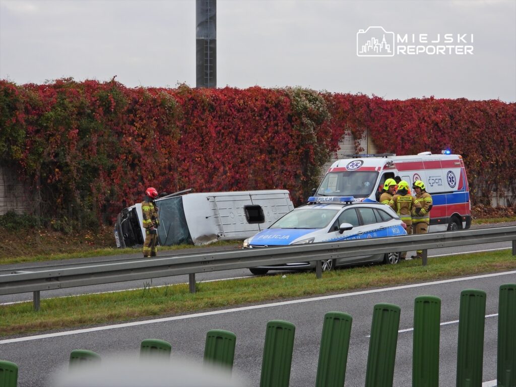 Ratownicy w kaskach rozmawiają obok przewróconego pojazdu na poboczu drogi, w tle widać ambulans i policyjny radiowóz.