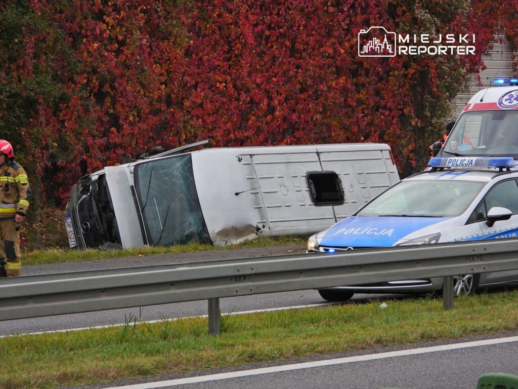 Na poboczu drogi leży przewrócony pojazd dostawczy, obok strażak w hełmie i radiowóz z włączonymi sygnałami świetlnymi.