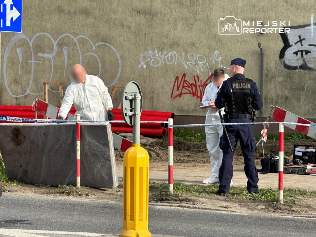 Mężczyzna w białym kombinezonie bada teren, podczas gdy policjant notuje informacje przy barierkach drogowych.