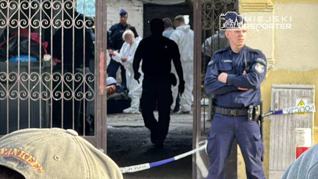 Grupa osób w białych kombinezonach bada miejsce zdarzenia, a policjant w mundurze obserwuje sytuację z boku.