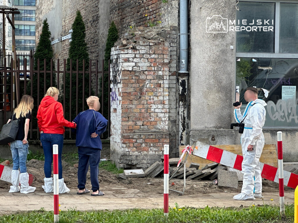 Grupa osób, w tym kobieta w czerwonej kurtce i dzieci, obserwuje mężczyznę w białym kombinezonie ochronnym, który dokumentuje miejsce zdarzenia.