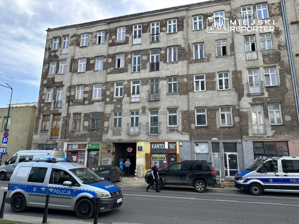 Policjanci rozmawiają z mężczyzną przed starym, zniszczonym budynkiem mieszkalnym.