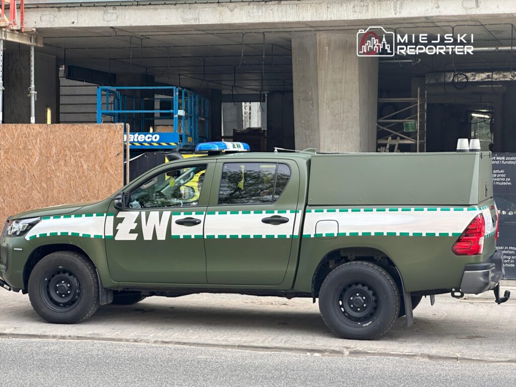 Samochód służbowy w zielonym kolorze z oznaczeniem "ZW" stoi na chodniku obok placu budowy.