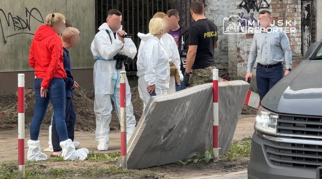 Grupa osób w odzieży ochronnej rozmawia z policjantem przy podwyższonej płycie betonowej w miejskim otoczeniu.