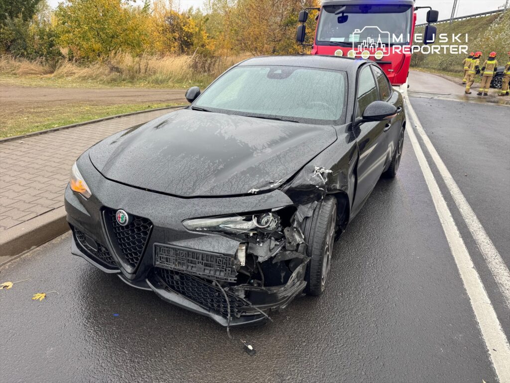 Czarny samochód osobowy z uszkodzoną przednią częścią stoi na drodze, obok widać czerwony wóz strażacki i ratowników.