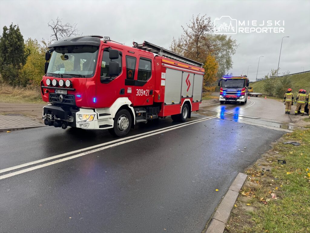 Czerwony wóz strażacki z włączonymi sygnałami świetlnymi stoi na drodze, obok drugiego wozu i strażaków w mundurach.