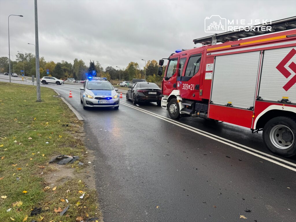 Policyjny radiowóz z włączonymi sygnałami świetlnymi stoi obok wozu strażackiego na drodze, w pobliżu miejsca wypadku.
