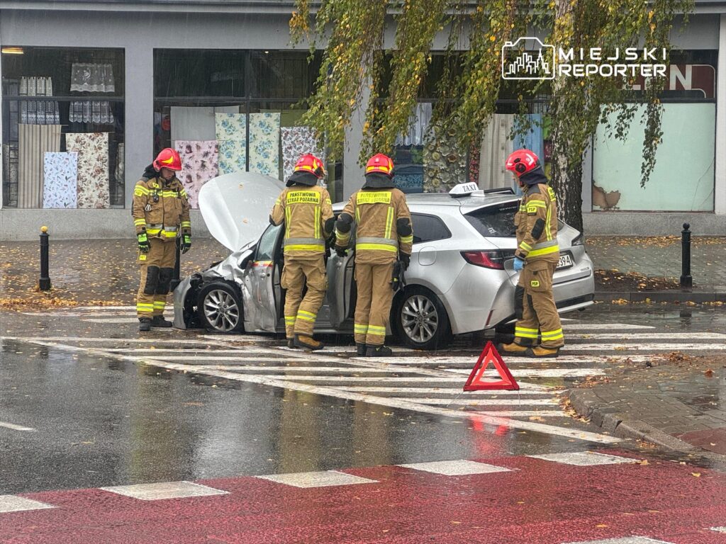 Czterech strażaków w mundurach interweniuje przy uszkodzonym samochodzie osobowym na deszczowej ulicy.