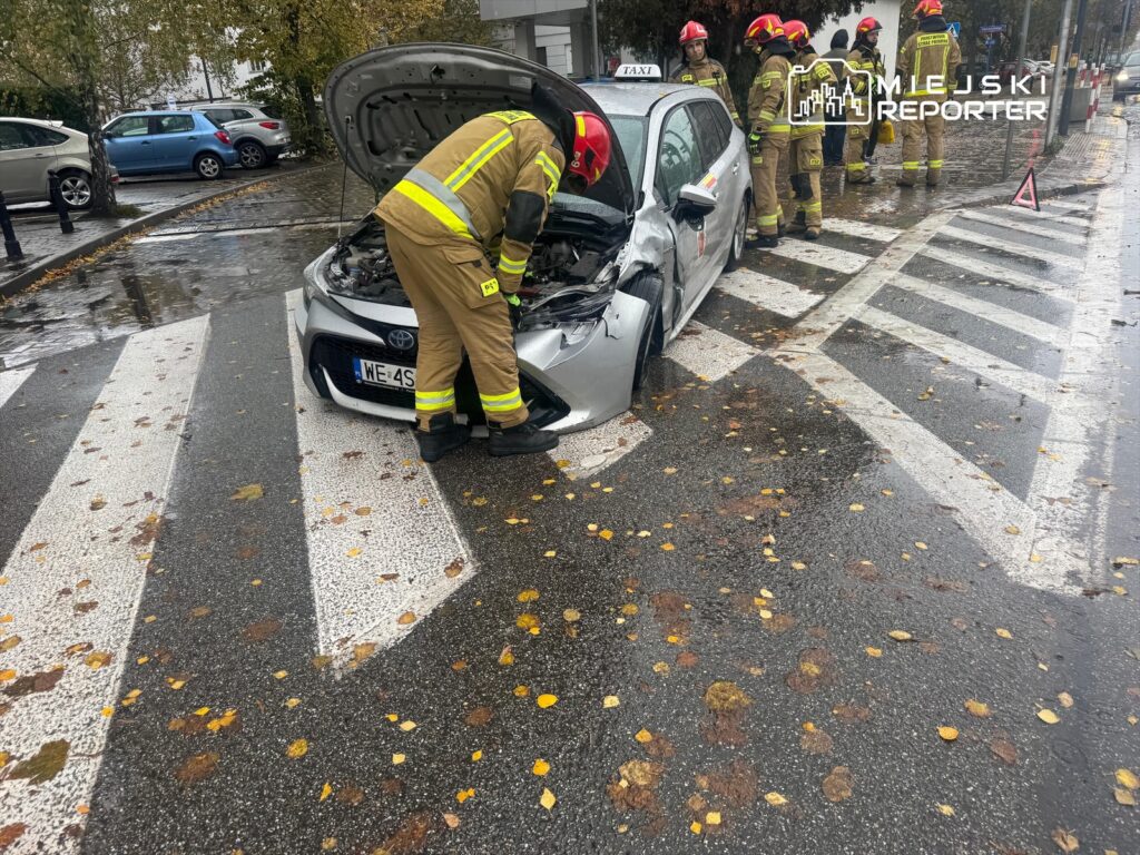 Strażacy sprawdzają uszkodzony samochód osobowy na pasach w deszczowej aurze, obok leżą liście.