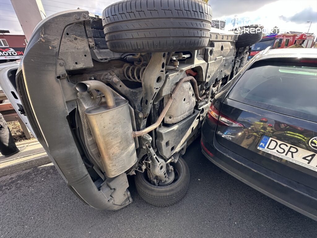 Przewrócony samochód osobowy leży na boku, z uszkodzoną karoserią, obok stoi drugi samochód.