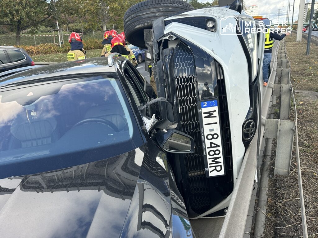 Odwrócone auto osobowe stoi na barierze drogowej, a w tle grupa strażaków w hełmach interweniuje.
