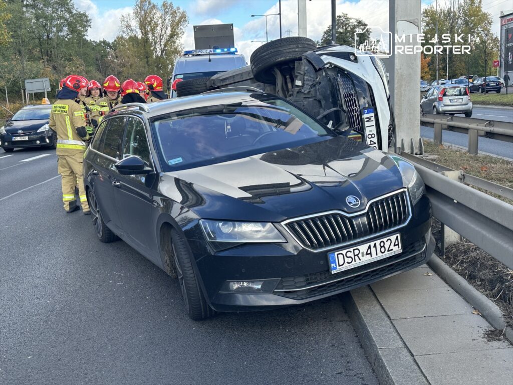 Grupa strażaków w hełmach omawia sytuację przy przewróconym samochodzie na poboczu drogi.