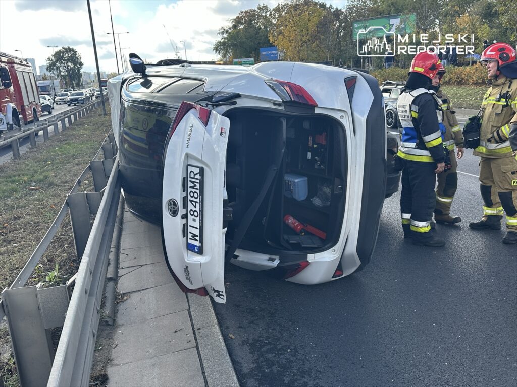Samochód osobowy leży na boku przy barierce, a obok trzech strażaków prowadzi rozmowę. W tle widać ruch uliczny.