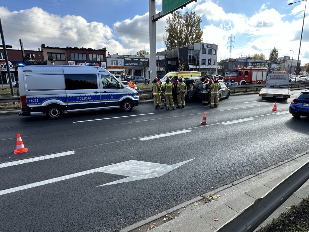 Na jezdni zatrzymane pojazdy służb ratunkowych, w tym radiowóz policji i wóz strażacki, otoczone przez ratowników.