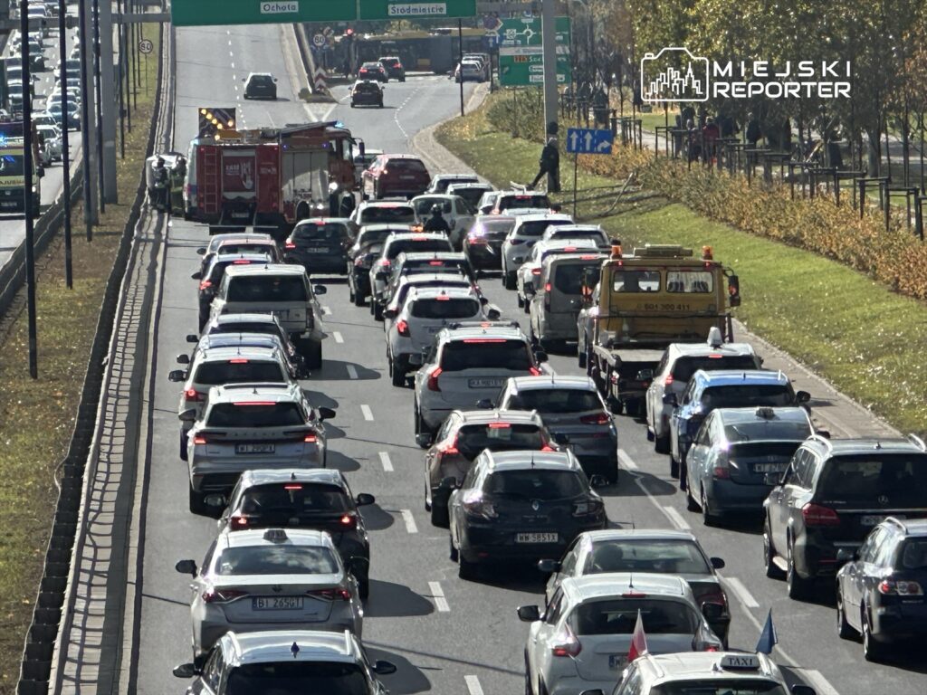 Ruch samochodowy w korku na drodze z włączonymi światłami awaryjnymi, obok stoi wóz strażacki.