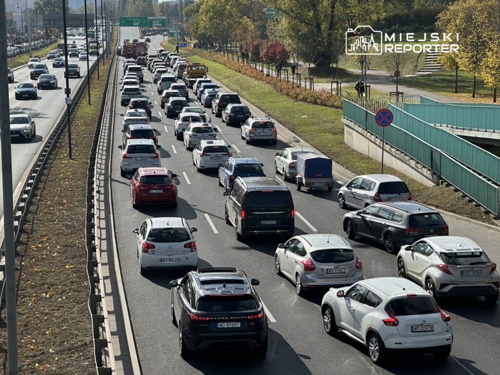 Zatłoczona droga z wieloma samochodami stojącymi w korku, w tle widoczne znaki drogowe i zielone drzewa.