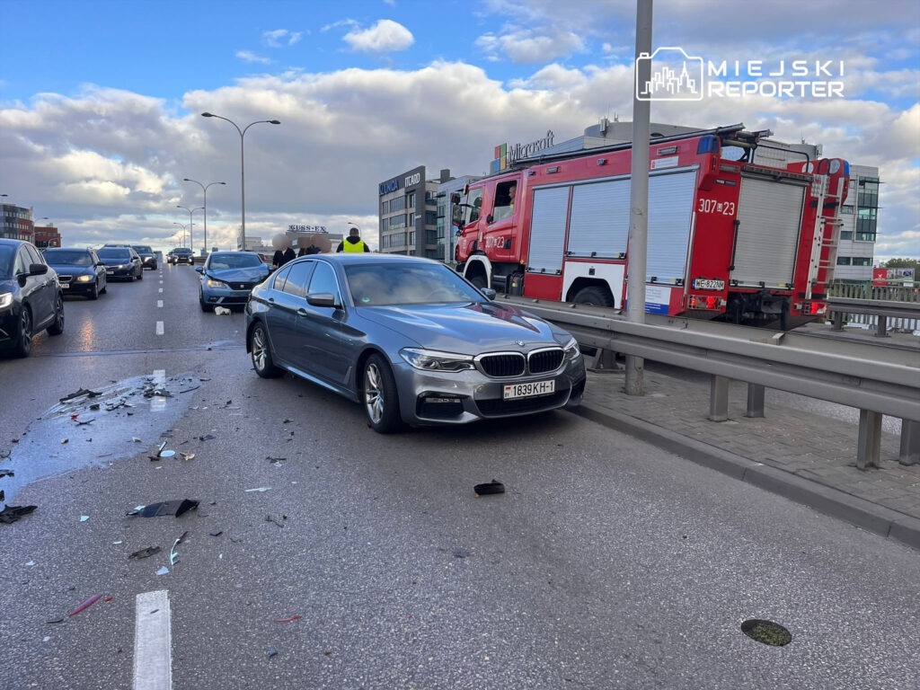 Kolizja drogowa z udziałem szarego BMW oraz uszkodzonymi pojazdami na ruchliwej ulicy, w tle wóz strażacki.