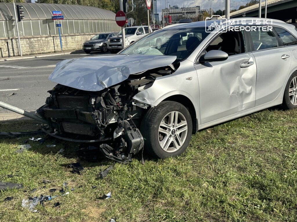 Uszkodzony samochód osobowy z wgniecionym przodem stoi na trawniku obok jezdni, otoczony fragmentami plastiku.