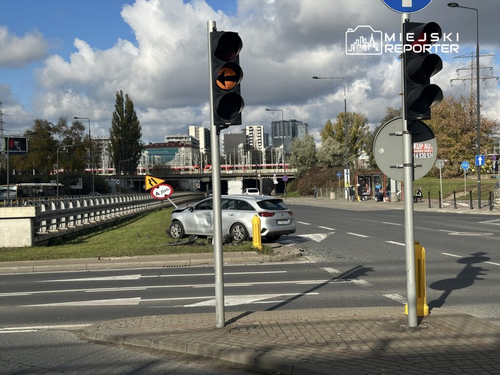 Samochód osobowy stoi na trawie obok skrzyżowania, z sygnalizacją świetlną w tle oraz znakiem drogowym.