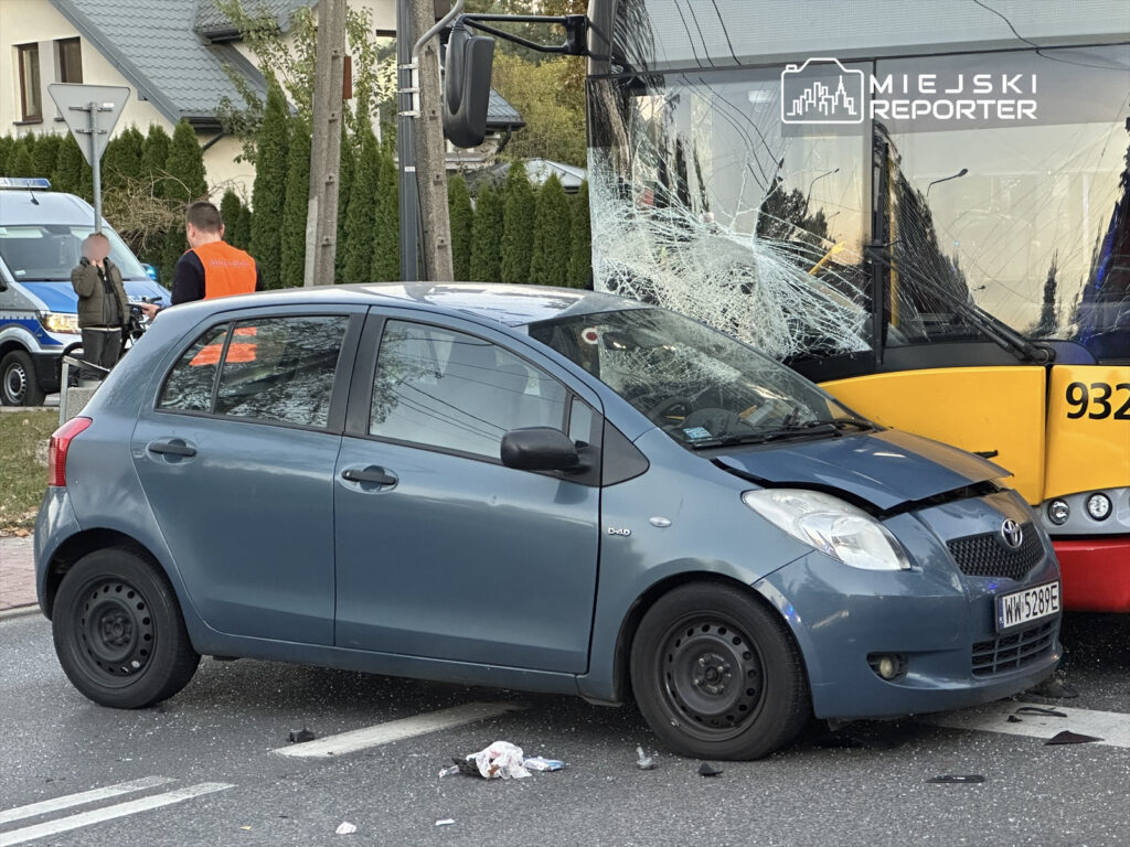 W miejscu wypadku stoi niebieski samochód osobowy z uszkodzoną maską obok autobusu z pękniętą szybą. Obok, mężczyzna w pomarańczowej kamizelce rozmawia z innym mężczyzną.