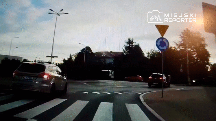 Samochód osobowy przejeżdża przez rondo na drodze z pasami, w tle widać budynki i sygnalizację drogową.