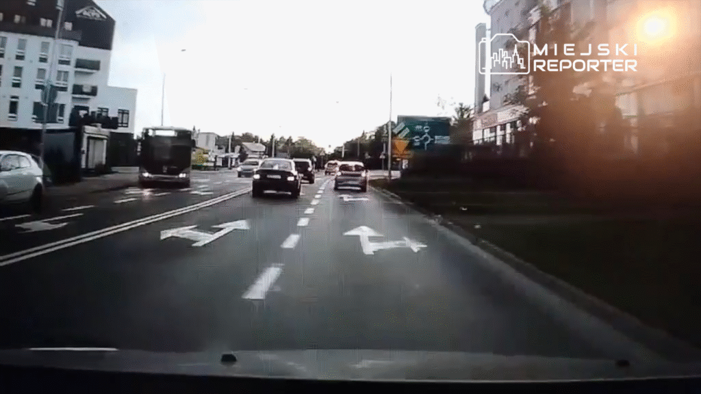 Ruchliwa ulica z samochodami poruszającymi się w różnych kierunkach, w tle nowoczesne budynki i znaki drogowe.