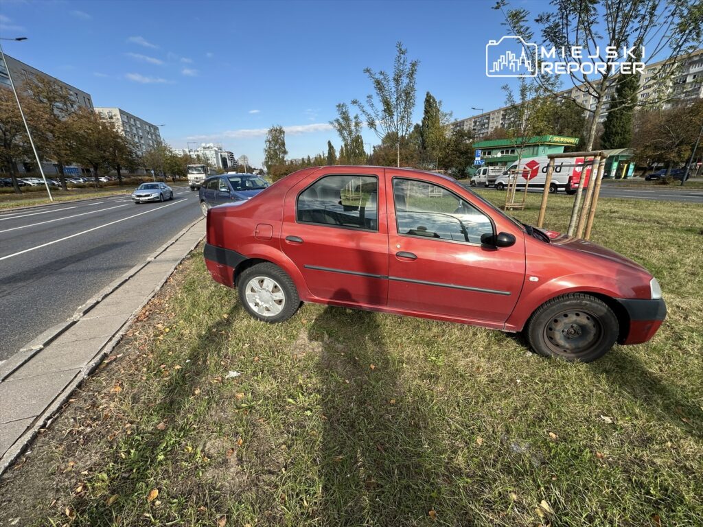 Czerwony samochód zaparkowany na trawie obok ruchliwej ulicy, w tle widoczne budynki mieszkalne.