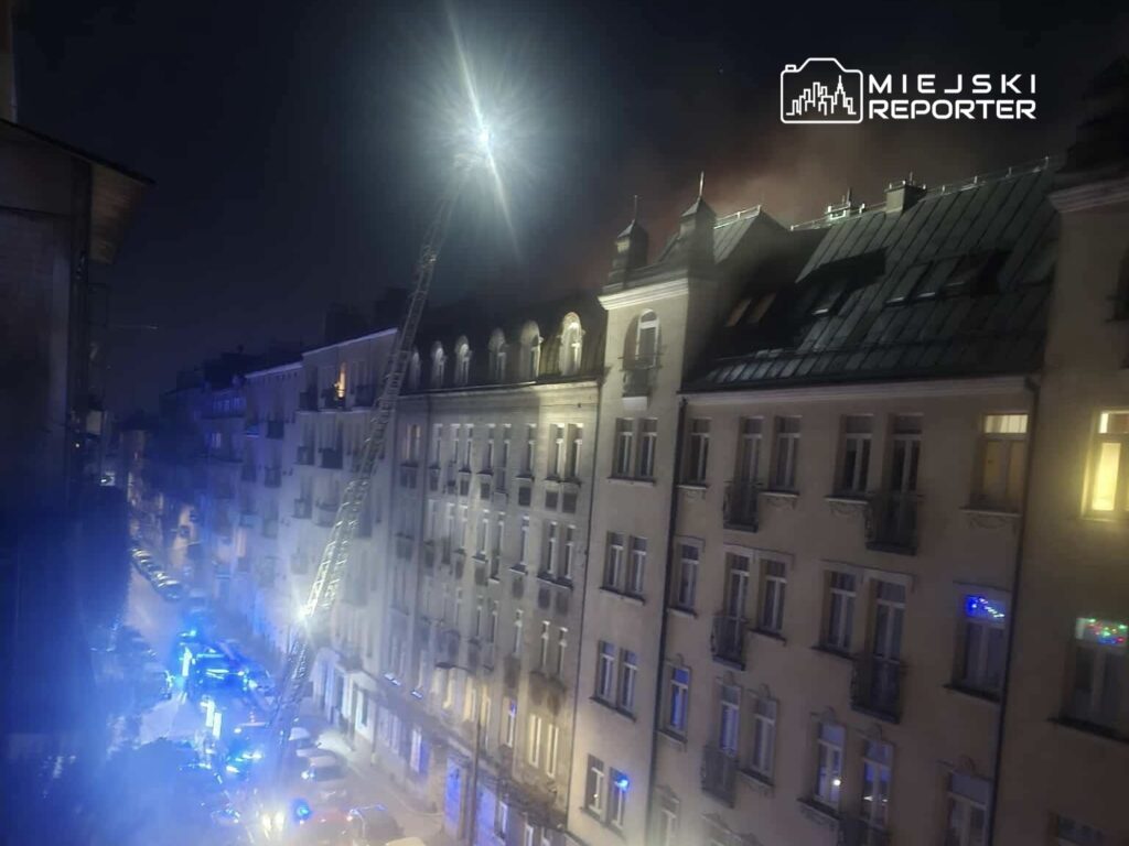 W nocy strażacy używają podnośnika do gaszenia pożaru na dachu budynku w miejskiej okolicy, otoczeni niebieskimi światłami.