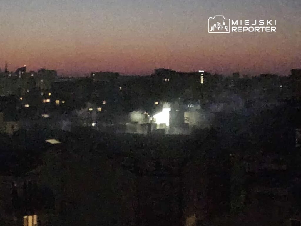 Widok na miasto o zmierzchu, z rozświetlonymi oknami budynków i delikatnym dymem unoszącym się w powietrzu.