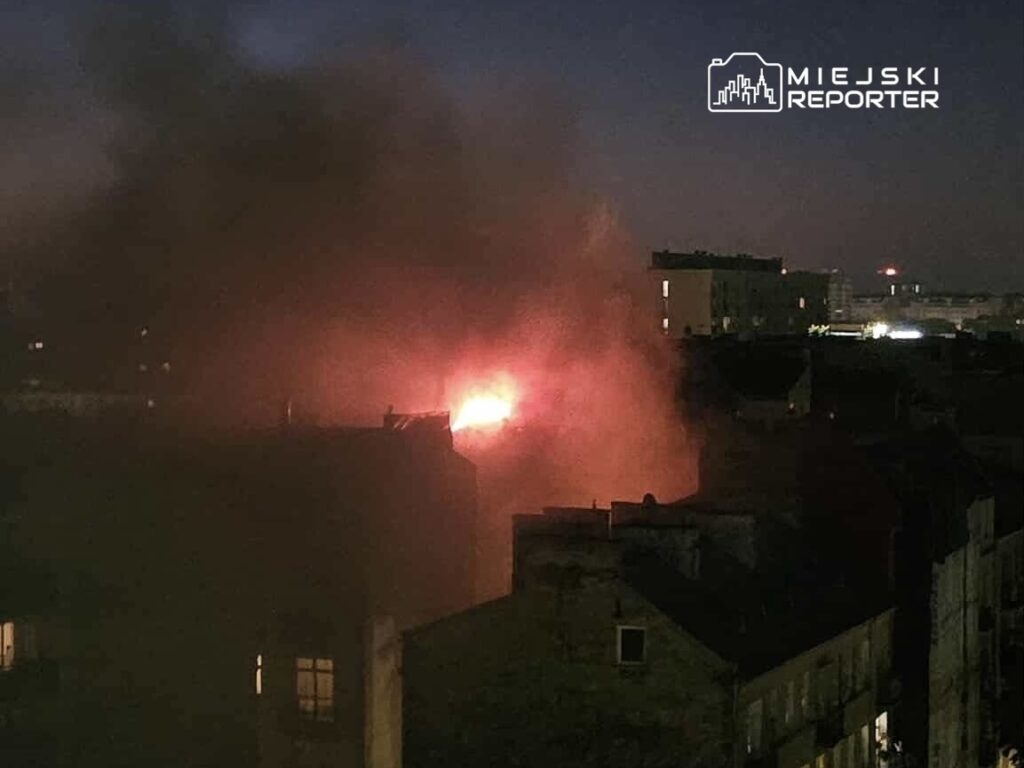 Płomienie i dym unoszą się nad budynkami w nocnej scenerii miejskiej.