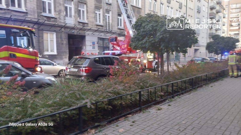 Strażacy przy wozie strażackim rozstawiają drabinę przed budynkiem mieszkalnym w miejskim otoczeniu.