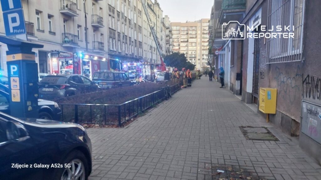 Grupa strażaków w odblaskowych kamizelkach pracuje przy wozie strażackim na ulicy, obok zaparkowanych samochodów.