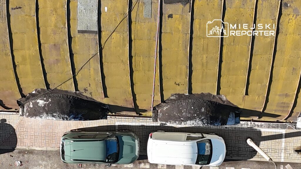 Dwa samochody zaparkowane na chodniku obok zniszczonego dachu z żółtej blachy.