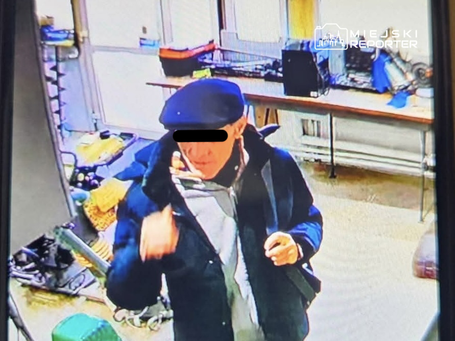 Mężczyzna w czapce i kurtce, gestykulujący w pomieszczeniu z narzędziami i meblami.