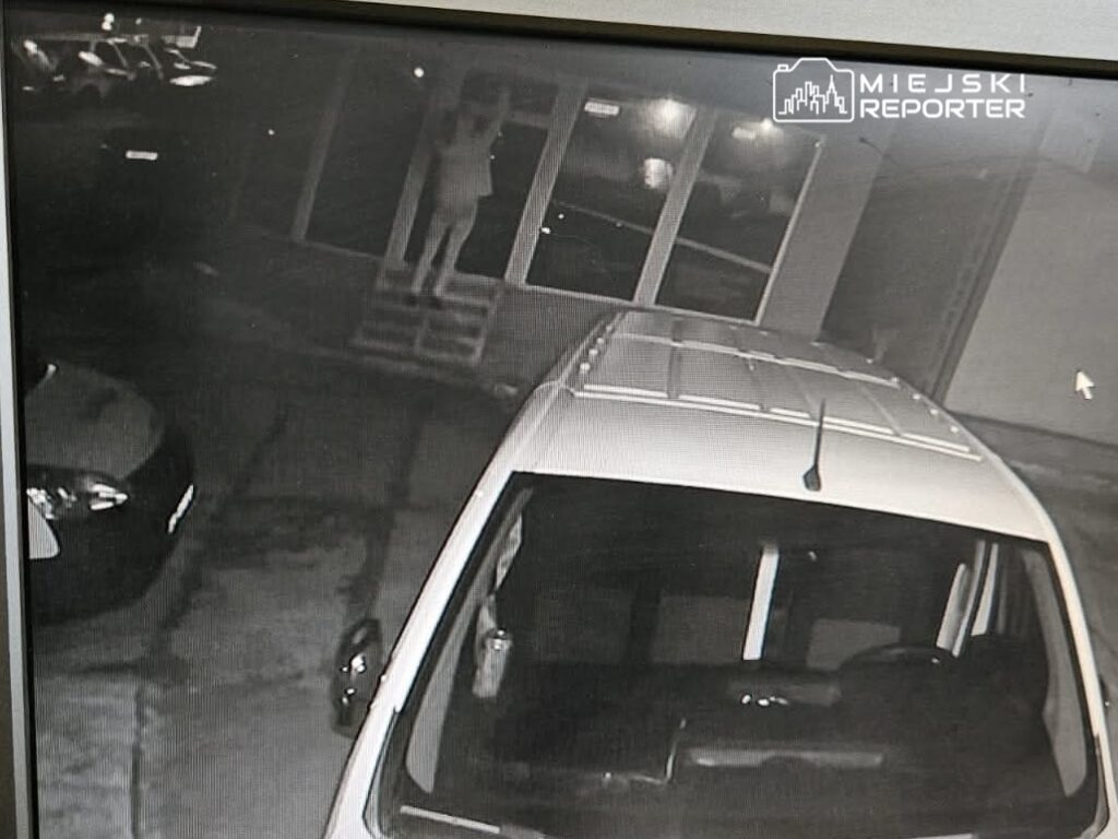 Osoba w jasnym ubraniu stoi na schodach przed wejściem do budynku, w tle widać zaparkowane samochody.
