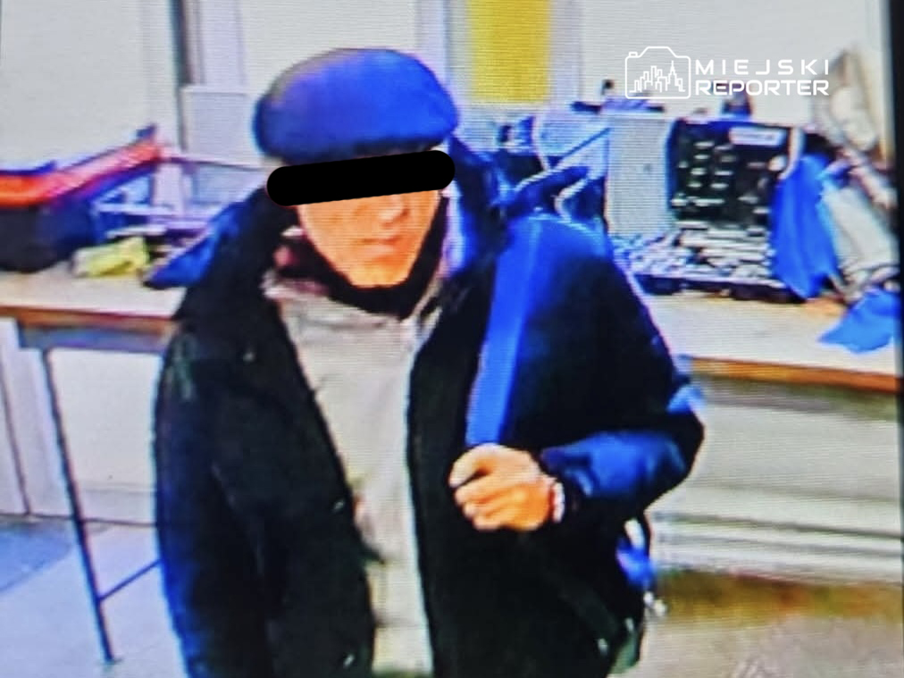 Mężczyzna w czapce i ciemnej kurtce trzyma torbę, stojąc w pomieszczeniu z komputerami i sprzętem elektronicznym.