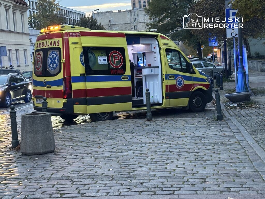 Ambulans z włączonymi światłami awaryjnymi zaparkowany na brukowanej ulicy, otwarte drzwi z widocznym wyposażeniem medycznym.