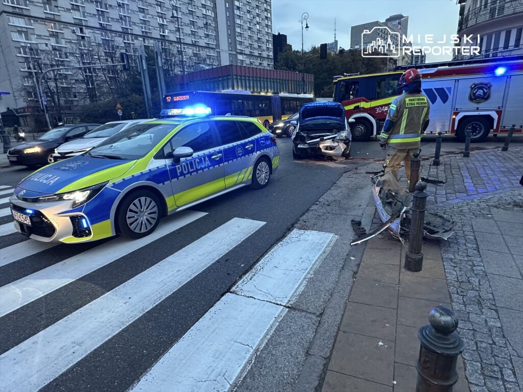 Policyjny radiowóz z niebieskimi światłami stoi na pasach, obok rozbite auto, a strażak ocenia sytuację.