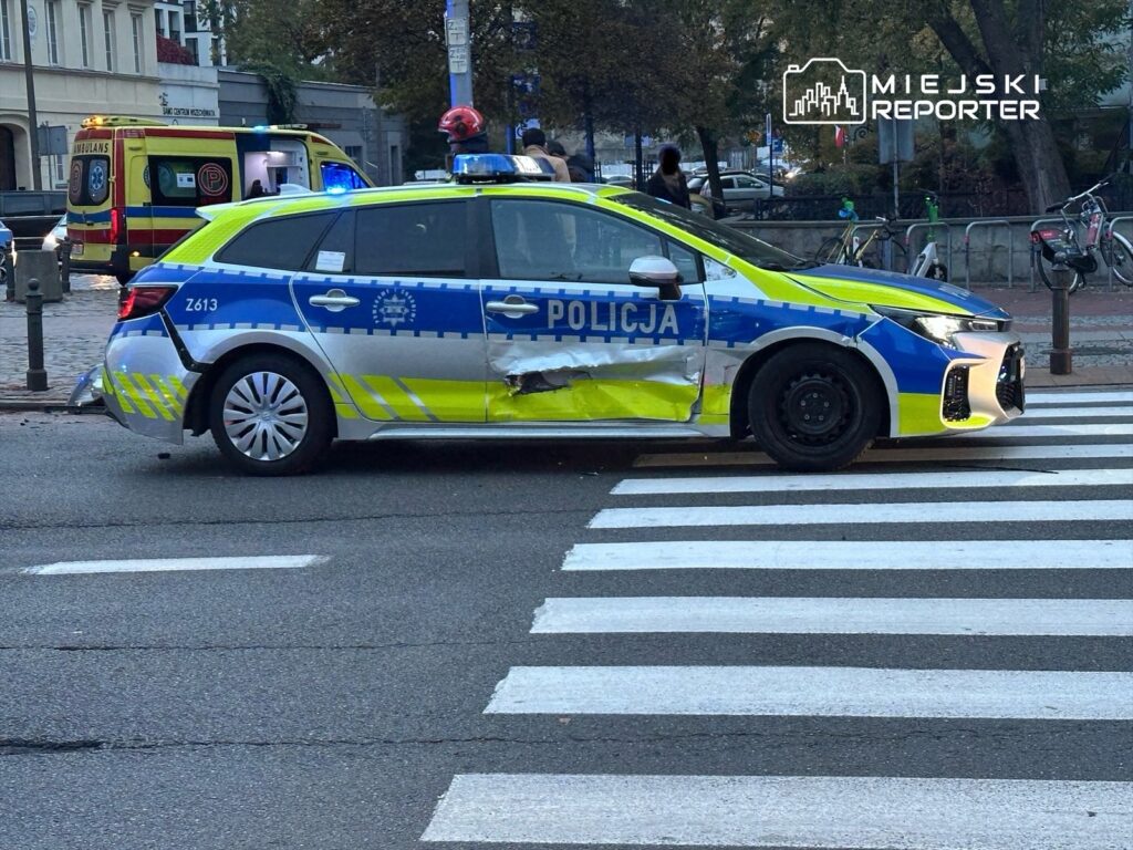 Policyjny samochód z uszkodzeniami stoi na przejściu dla pieszych, w tle widać ambulans.