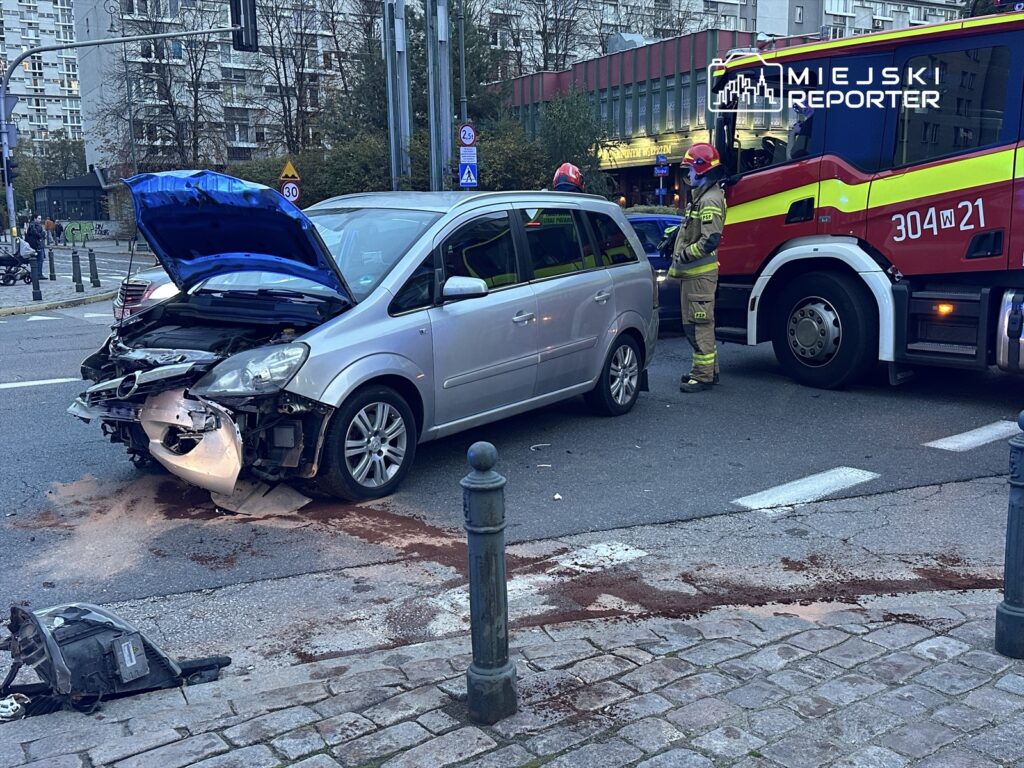 Uszkodzony samochód osobowy z otwartą maską stoi na ulicy, obok strażak rozmawia z kierowcą. W tle widoczne budynki.