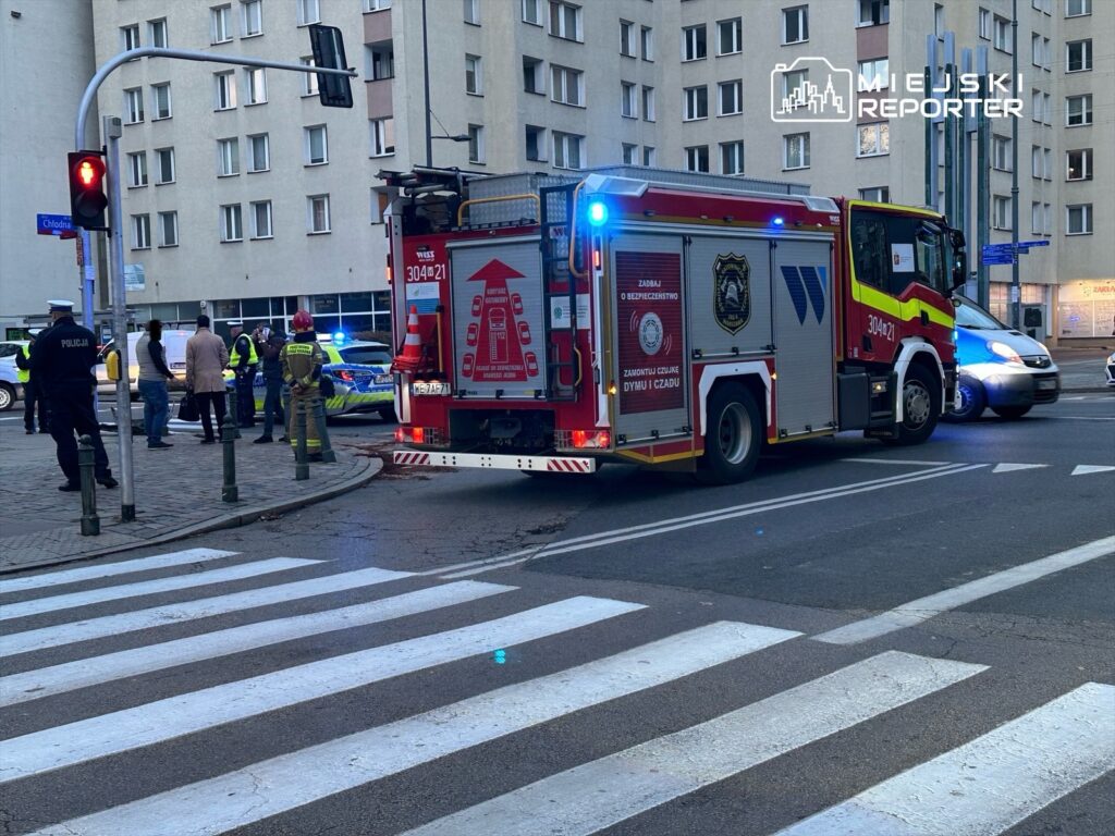 Straż pożarna z włączonymi sygnałami świetlnymi zatrzymuje się przy ulicy, a policja zabezpiecza miejsce zdarzenia.