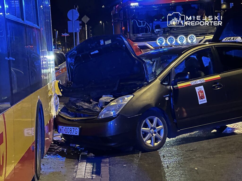 Zderzenie taksówki z autobusem miejskim, z otwartą maską pojazdu i widocznymi uszkodzeniami. Nocna sceneria z niebieskimi światłami.