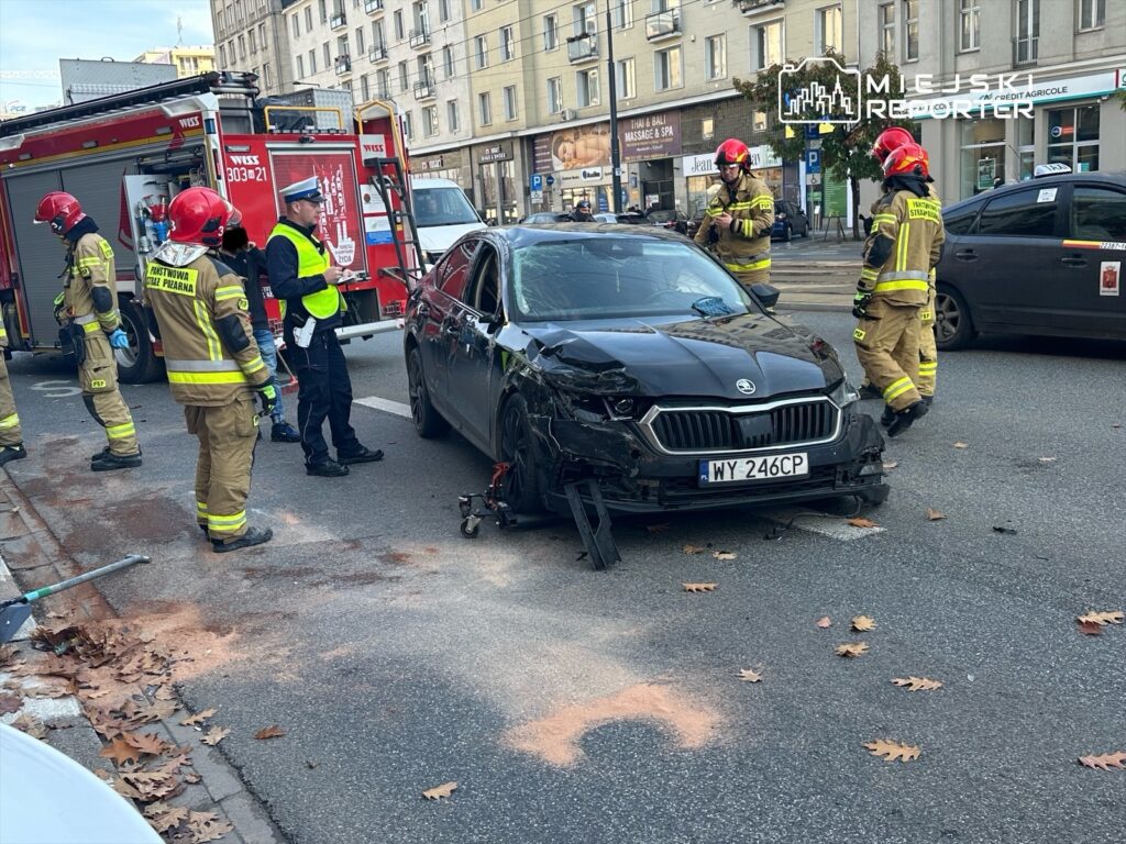 Grupa strażaków i policjantów ocenia uszkodzone czarne auto na ulicy, obok wozu strażackiego. Na jezdni widoczne liście.