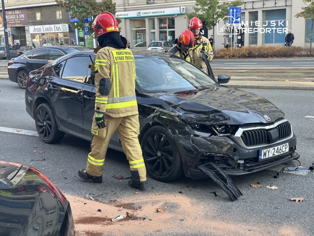 Strażacy w żółtych mundurach sprawdzają uszkodzone czarne auto osobowe na ulicy, obok leżą resztki piasku.