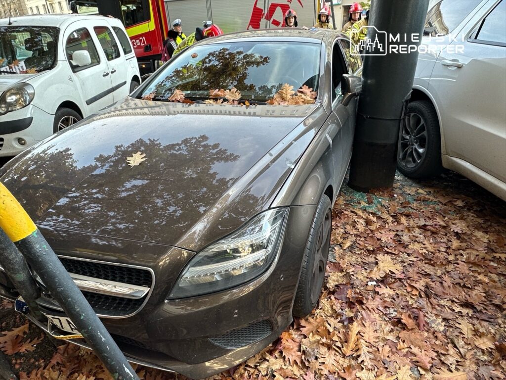 Samochód osobowy uszkodzony na parkingu, zderzony z metalowym słupem, otoczony opadłymi liśćmi.