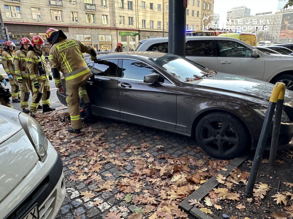 Strażacy w mundurach, otwierający drzwi ciemnoszarego samochodu osobowego na ulicy pokrytej opadłymi liśćmi.