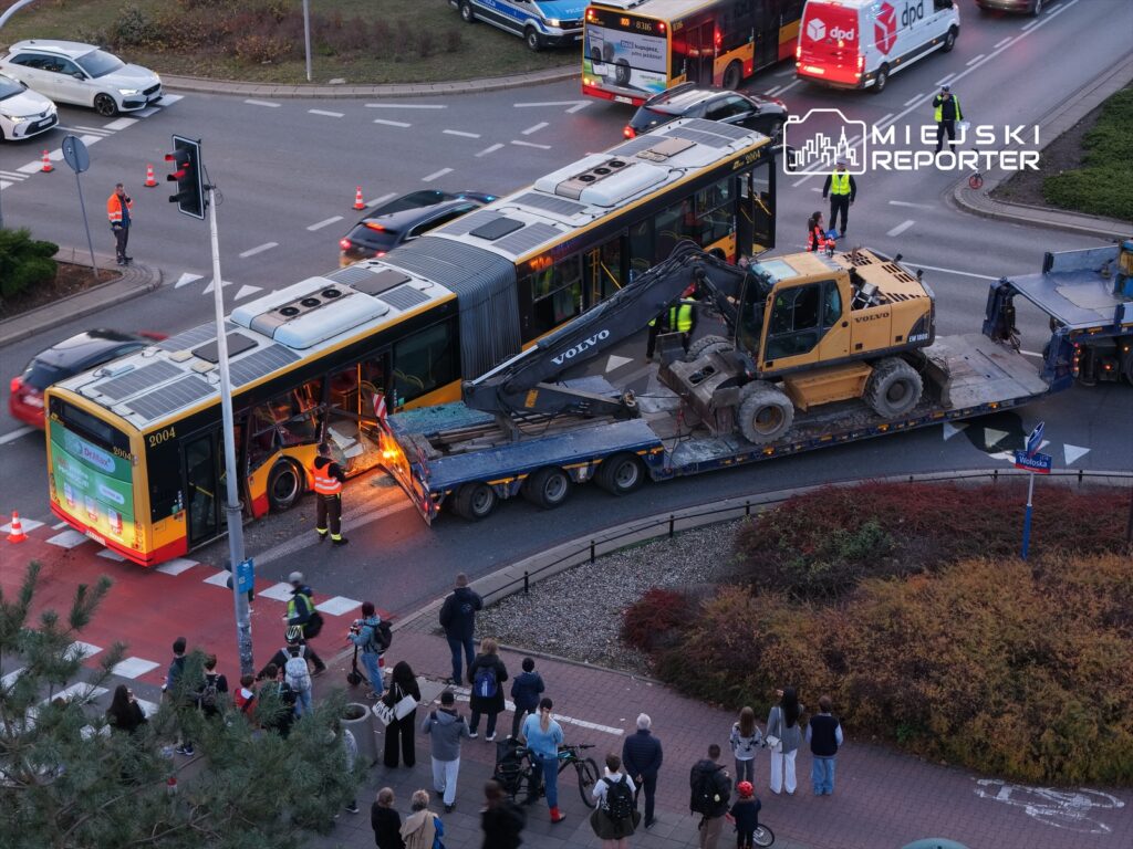 Autobus z uszkodzoną tylną częścią jest holowany przez ciężki sprzęt, a obok gromadzi się grupa gapiów.